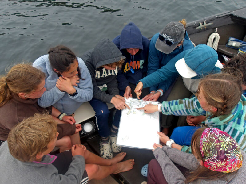 A group of people are gathered in a boat, looking at a map or chart. They are pointing and discussing something, likely planning a route or analyzing data. The atmosphere seems collaborative and focused, suggesting a shared activity or project. The setting appears to be outdoors, possibly on a lake or river.